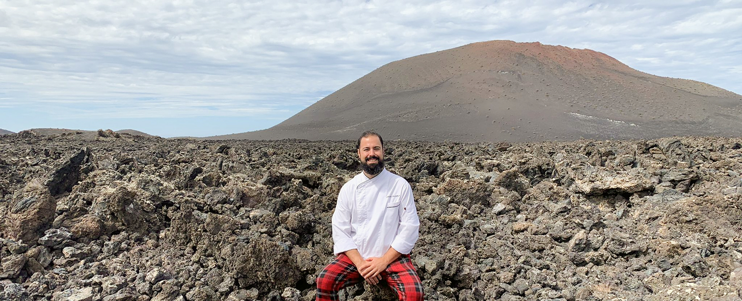 El chef Gonzalo Calzadilla, con chaquetilla blanca y pantalón de cuadros rojos, posa sonriente sobre el campo de lava del Parque Nacional de Timanfaya, Lanzarote, con un volcán al fondo.