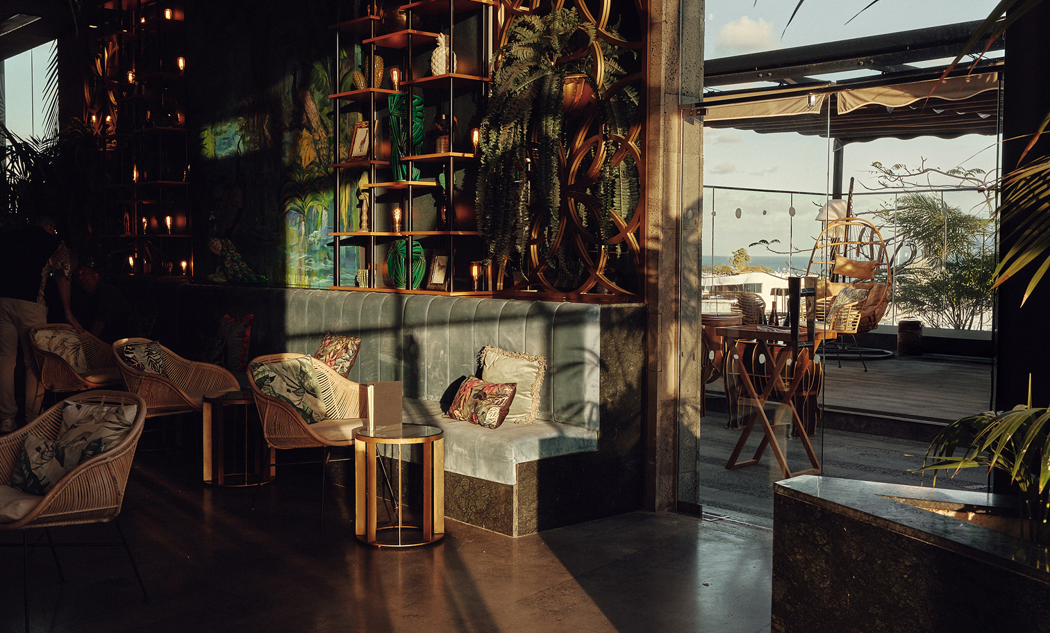 Interior de restaurante en Lanzarote con decoración tropical y luz cálida. Banqueta tapizada, sillones de ratán y estanterías con plantas y bombillas Edison crean el escenario perfecto para una cena a 4 manos. Al fondo, terraza con vistas al exterior.