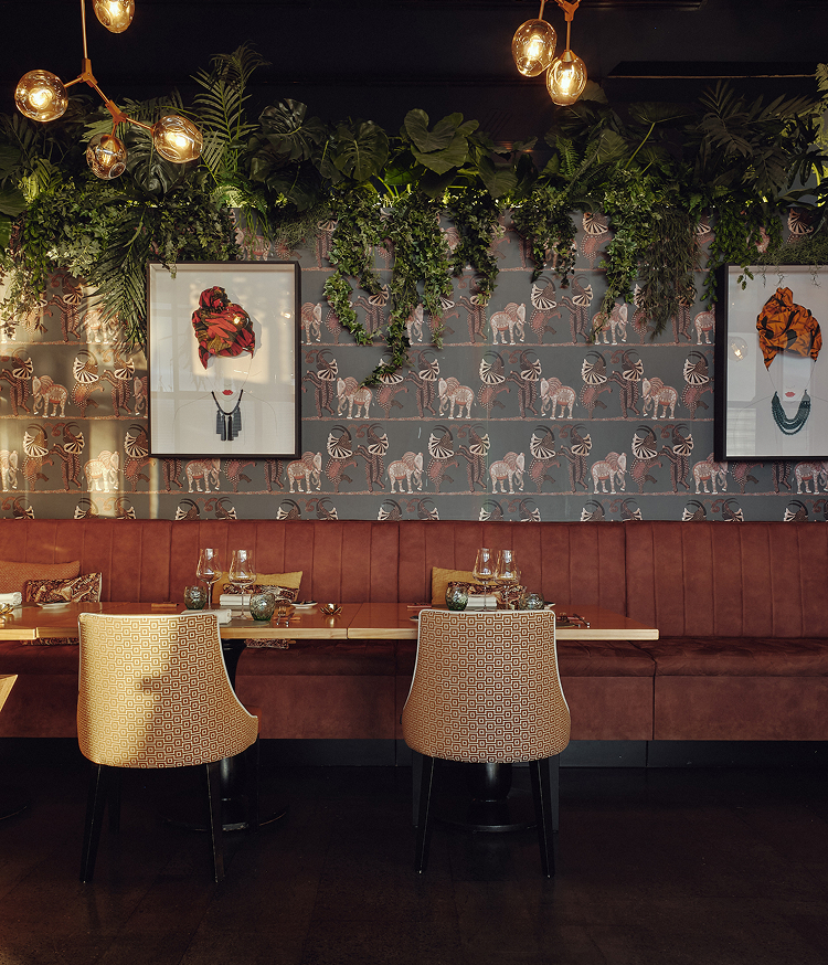 Sala interior de restaurante en Lanzarote con banqueta de terciopelo burdeos, sillas tapizadas en dorado y mesas preparadas para una cena a 4 manos. Papel pintado con motivos de elefantes, cuadros de arte contemporáneo y jardín colgante con luces doradas completan el ambiente.