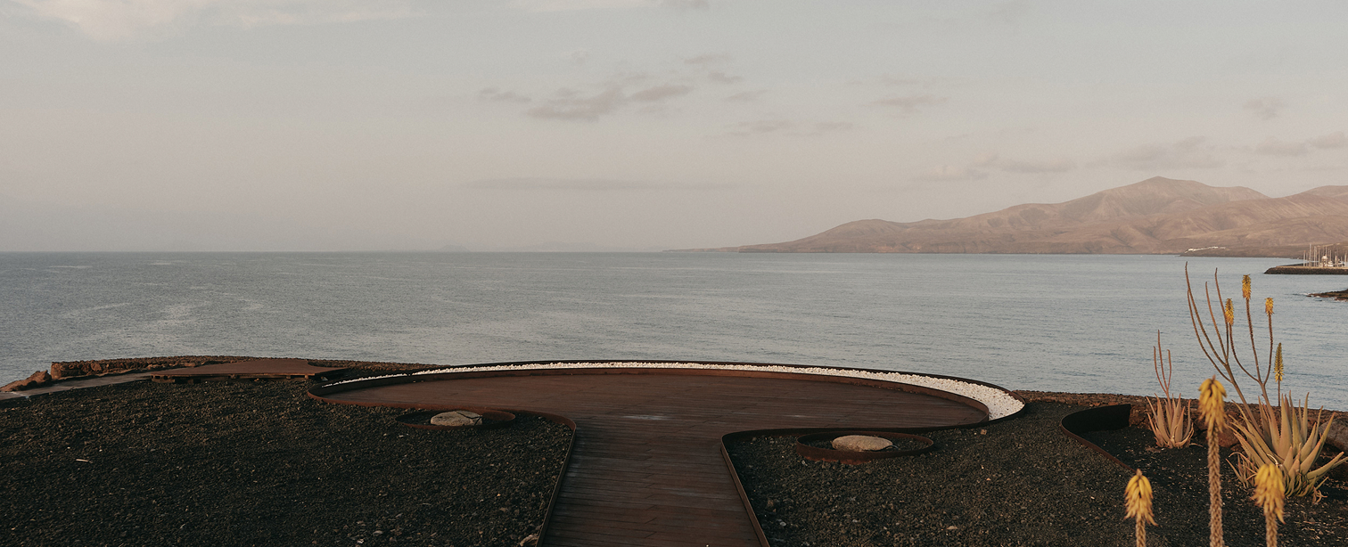Mirador de madera con forma orgánica en la costa de Lanzarote al atardecer. Una pasarela conduce a una plataforma circular sobre suelo de picón volcánico, con vistas al océano Atlántico y una montaña al fondo. A la derecha, plantas de aloe vera en flor.