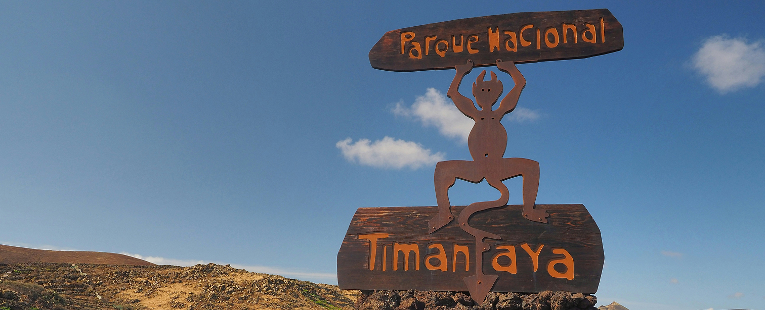 Cartel de entrada al Parque Nacional de Timanfaya, Lanzarote. Una figura metálica del diablo —mascota del parque— sostiene sobre su cabeza un tablón con el texto 