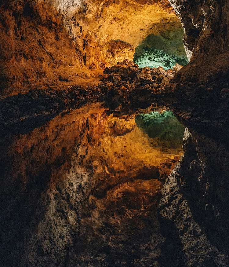 Interior de la Cueva de los Verdes, Lanzarote. Túnel volcánico iluminado con tonos dorados y verdes que se refleja simétricamente en un lago subterráneo de aguas quietas.
