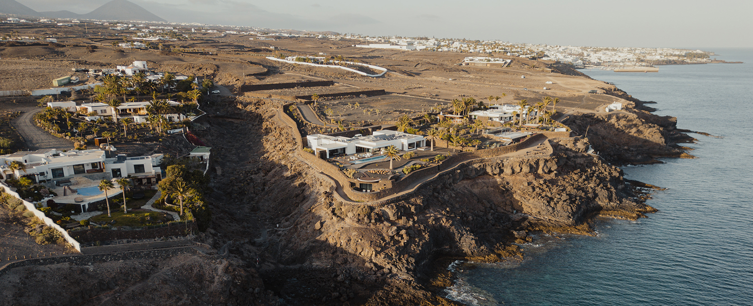 Vista aérea de una urbanización costera en Lanzarote. Villas blancas de arquitectura moderna con piscinas se asientan sobre acantilados volcánicos rocosos frente al océano Atlántico. Palmeras y vegetación xerófila salpican el paisaje árido. Al fondo, el núcleo urbano blanco de Playa Blanca y volcanes en el horizonte.