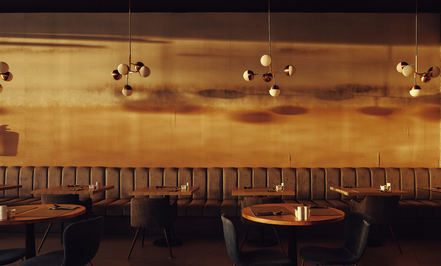 Interior de restaurante de diseño con iluminación cálida. Banqueta corrida tapizada en terciopelo gris frente a mesas redondas de madera con cubiertos y velas. Lámparas colgantes de latón con esferas iluminan una gran pared dorada que refleja los juegos de luz. Ambiente íntimo y sofisticado.