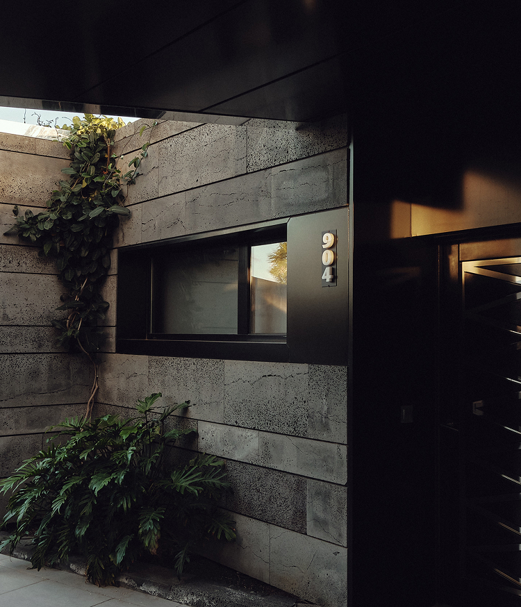 Fachada de arquitectura contemporánea con revestimiento de piedra volcánica oscura en hiladas horizontales. Una ventana de marco negro muestra el número 904. Plantas trepadoras y vegetación tropical enmarcan la entrada, junto a una puerta de lamas metálicas con luz cálida interior.